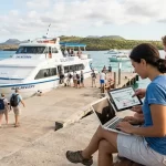A couple sits on a wooden bench at the harbor, focused on their laptop and tablet to securely check Galaferry ticket prices for their upcoming inter-island trip.