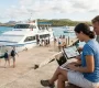 A couple sits on a wooden bench at the harbor, focused on their laptop and tablet to securely check Galaferry ticket prices for their upcoming inter-island trip.